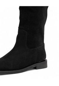 Stivale nero in suede alta fino al ginocchio con un taglio laterale rettangolare, texture liscia e tacco basso a blocco. Dettagli di cucitura minimi.