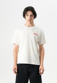 Cream-colored cotton t-shirt with short sleeves and a round neckline, featuring a small red graphic print on the chest. Dark denim jeans.
