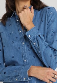 Lichtblauw denim shirt met geborduurde kleine hartjes in donkerblauw. Knopen aan de voorkant en bij de manchetten. Lange mouwen.