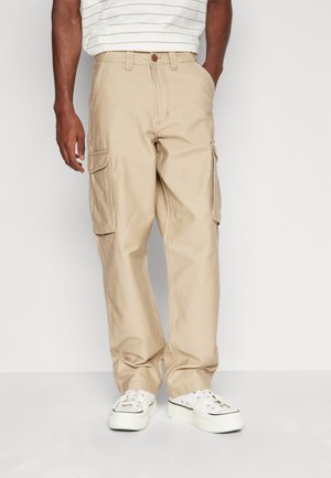 Beige Cargo-Hosen mit Seitentaschen, Knopfverschluss und lockerer Passform. Kombiniert mit weißen Sneakers und einem gestreiften T-Shirt.