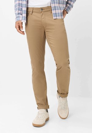 Mand iført opkørte beige bukser, hvide sneakers, en hvid skjorte og en blå og lyserød ternet langærmet skjorte.