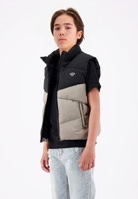 Geplüschte ärmellose Weste in Schwarz und Hellgrau, mit hohem Kragen, vorderem Reißverschluss und Seitentaschen, sowie einem Logo auf der Brust.