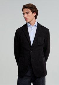 Blazer negro texturizado con un solo bolsillo en el pecho, solapas con muesca y dos bolsillos frontales, combinado con una camisa de color azul claro.