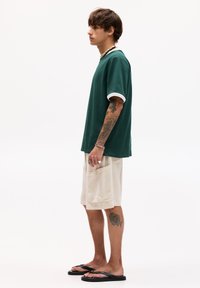 T-shirt en coton vert avec des finitions blanches, associé à un short beige texturé. Sandales noires portées, avec un tatouage sur la jambe inférieure.