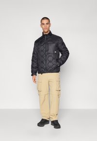 Sort, quiltet pufferjakke med hexagonalt mønster, høj krave og lynlås foran. Sammen med beige cargo-bukser og sorte sko.