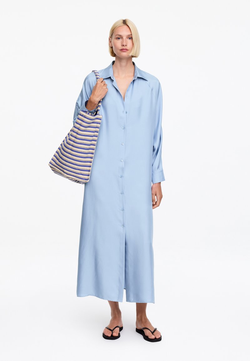Femme aux cheveux blonds courts portant une longue robe bleu clair boutonnée, portant un sac fourre-tout rayé bleu et beige, et des tongs noires.