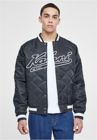 Black quilted bomber jacket with white accents, featuring a logo on the chest, snap front closure, and striped ribbed cuffs and hem.