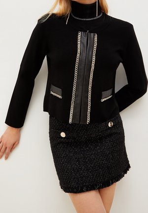 Kvinde bærer sort langærmet top med læder- og kædebesætning, kombineret med en tekstureret sort mininederdel med metalliske knapper og frynsekant.