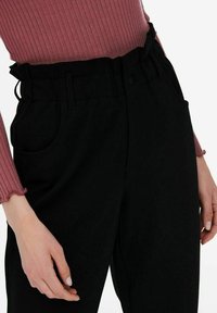 Zwarte broek met hoge taille en een elastische tailleband, zijzakken en een gladde, matte stof. Gekleed met een geribbeld roze top.
