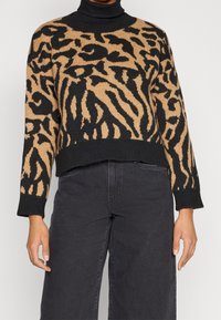Eine Frau trägt einen verkürzten Pullover mit Tiermuster in Schwarz und Beige über einem schwarzen Rollkragenpullover, kombiniert mit hochtaillierten schwarzen weiten Hosen.