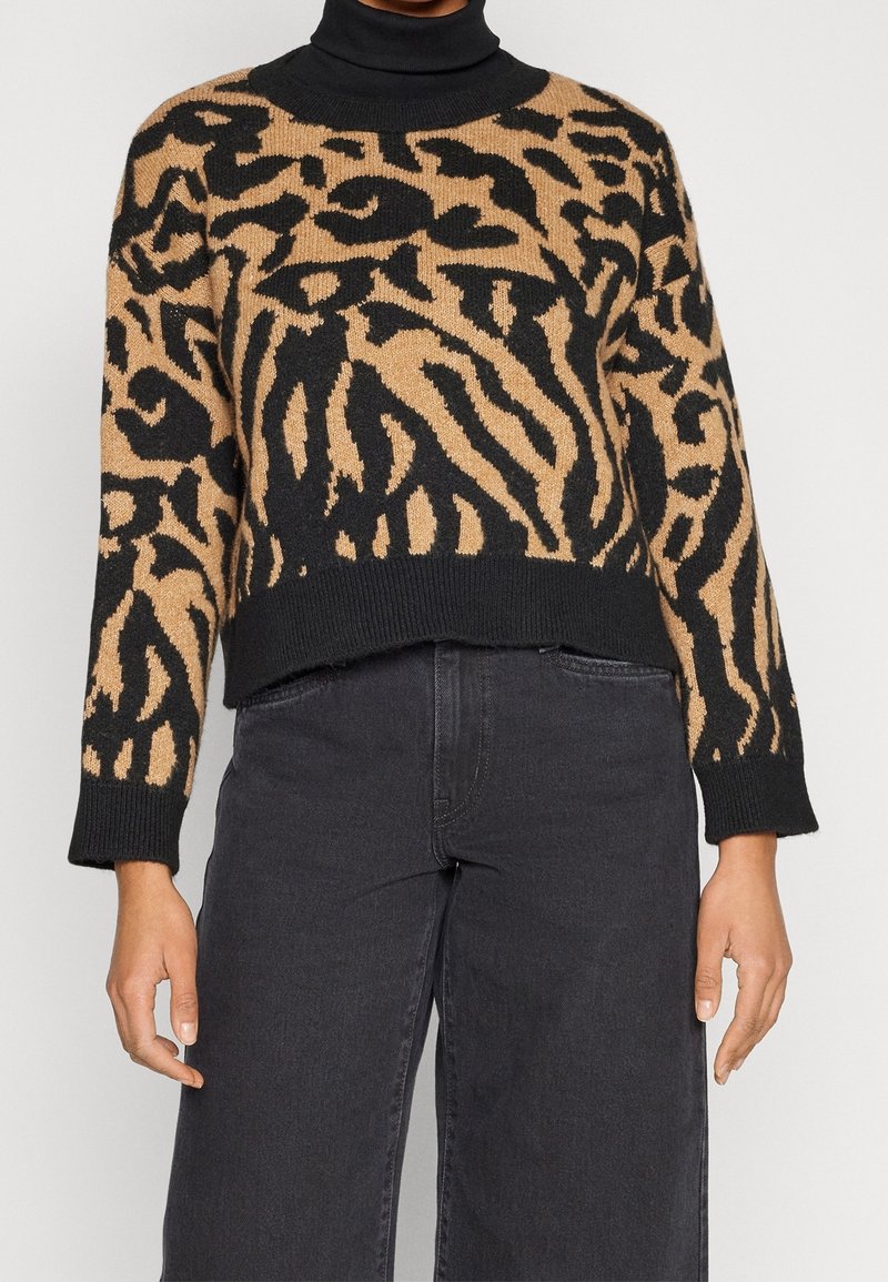 Eine Frau trägt einen verkürzten Pullover mit Tiermuster in Schwarz und Beige über einem schwarzen Rollkragenpullover, kombiniert mit hochtaillierten schwarzen weiten Hosen.