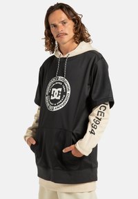 Felpa nera e beige con un grande logo circolare sul davanti, vestibilità oversize, maniche corte e tasca frontale. Presenta un cappuccio con cordini.