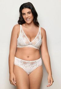 Uśmiechnięta kobieta z ciemnymi włosami, ubrana w pasujący biały koronkowy biustonosz i figi, stojąca na tle jasnego, jednolitego tła.