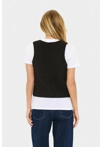 Vrouw met lang blond haar die een zwart gebreid vest draagt over een wit T-shirt met korte mouwen en donkerblauwe jeans, met haar rug naar de camera gekeerd tegen een witte achtergrond.