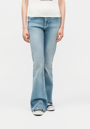 Άτομο που φορά αποκλειστικά ανοιχτό μπλε τζιν με φαρδιά μπατζάκια και μαύρα-άσπρα αθλητικά παπούτσια, στέκεται μπροστά σε ένα απλό λευκό φόντο.