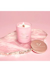 Różowa świeca w szklanym pojemniku z złotymi akcentami, zapalony knot i detal z kryształowym logo. Umieszczona na podstawie z jasnoróżowego agatu.