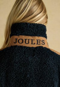 Kurtka z sztucznego futra o granatowej fakturze, z brązowym panelem na ramionach i haftowanym napisem "JOULES" na kołnierzu. Staranny, przytulny design.