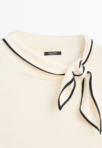 Pull crème avec un col bordé de noir et un détail de nœud en tissu assorti bordé de noir sur le côté droit.