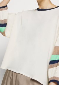 Vit topp med ribbad, flerfärgad randig ärmar i beige, blått och grönt. Har en rund halsringning med en mörkblå kant.