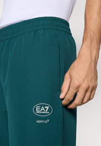 Záber zblízka osoby nosiacej elastické nohavice s rozšíreným pásom v tyrkysovej farbe s logom "EA7 Emporio Armani VENTUS7" blízko vrecka, ruka odpočívajúca vedľa.