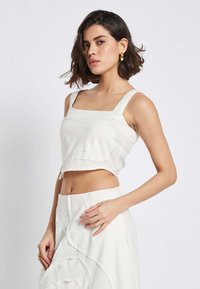 Weißes Cropped-Top mit quadratischem Ausschnitt und breiten Trägern. Mit fransigen Kanten und strukturierten Details. Kombiniert mit einem passenden, hoch taillierten Rock.