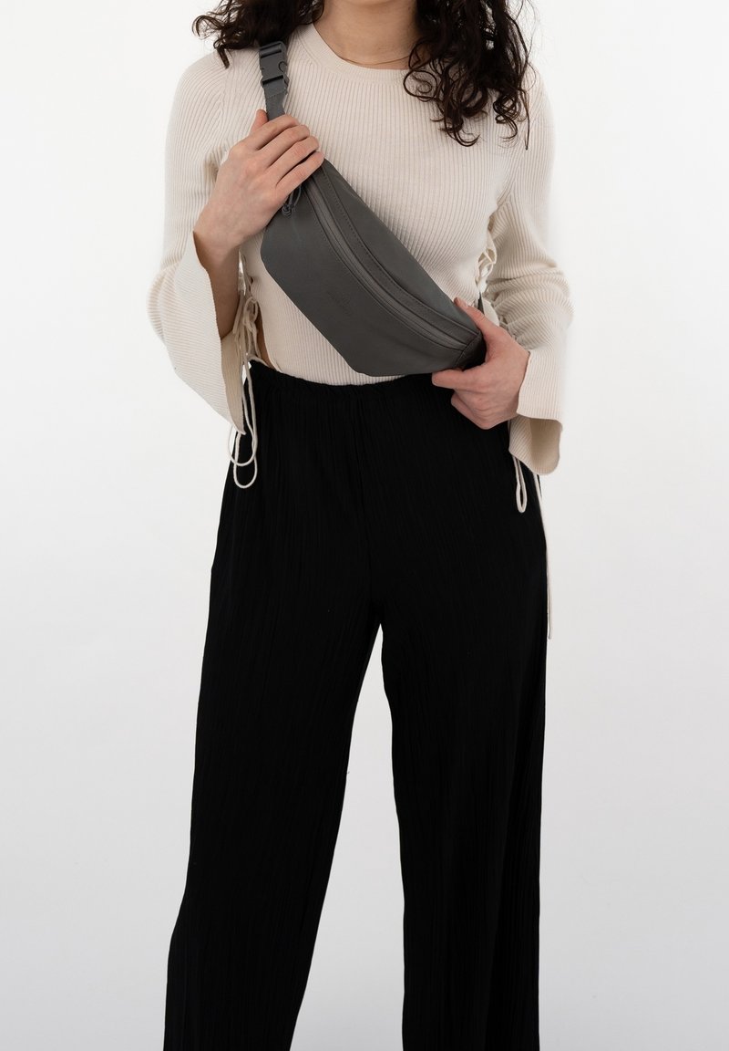 Femme portant un pull côtelé crème et un pantalon noir, tenant un sac gris porté en bandoulière sur la poitrine sur fond blanc.