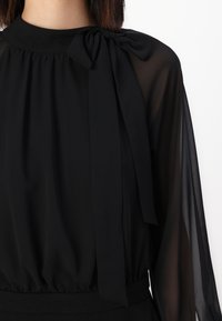 Zwarte blouse met hoge hals, strikaccent en doorschijnende lange mouwen. Elastische taille en een gladde textuur. Elegante stijl geschikt voor formele gelegenheden.