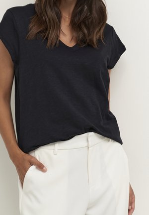 Femme portant un haut noir à manches courtes en V rentré dans un pantalon blanc taille haute, une main dans une poche.