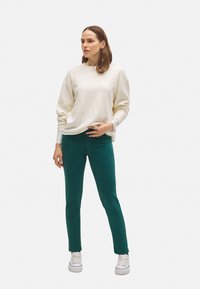 Sudadera blanca de felpa con hombros caídos, combinada con pantalones ajustados verdes y zapatillas blancas. Texturas suaves y un diseño casual.