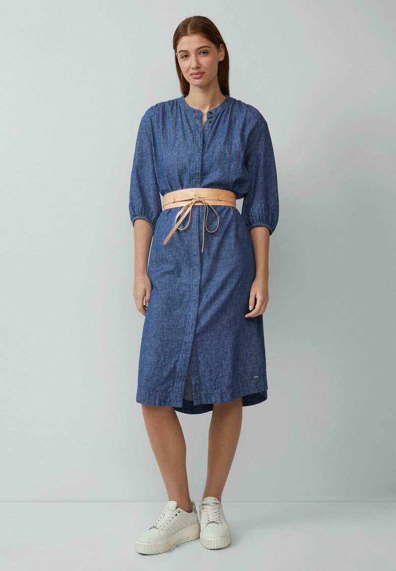 Jeans-Midi-Kleid in Blau mit durchgehendem Knopfverschluss, dreiviertel Ärmeln und einem beigen Gürtel. Kombiniert mit weißen Plateausneakern.