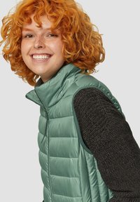 Hellgrüne gepolsterte Weste mit hohem Kragen, Reißverschluss vorne und schwarz strukturierten Strickärmeln. Weicher Stoff, leichtes Design.