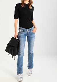 Top negro de manga corta con textura acanalada combinado con jeans de pierna recta en azul claro con cinco bolsillos, zapatillas plateadas y una mochila negra.