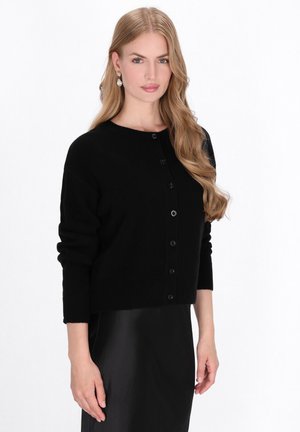 Schwarzer gestrickter Cardigan mit rundem Halsausschnitt, Knopfverschluss und gerippten Bündchen. Aus weichem Stoff, über einem schwarzen Rock getragen.