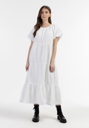 Femme aux longs cheveux bruns portant une robe midi blanche à volants et des bottes de combat noires à lacets, debout sur un fond blanc.