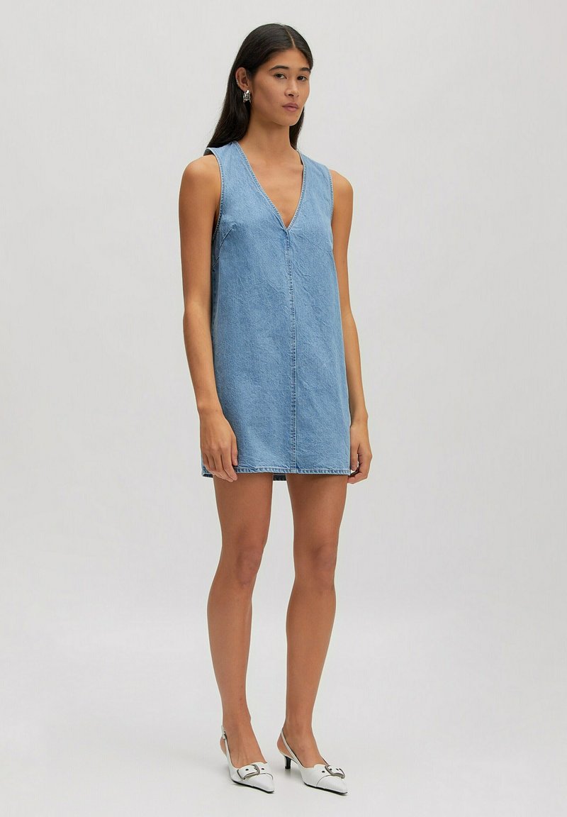 Vrouw met lang donker haar, gekleed in een blauwe mouwloze denim mini-jurk en witte puntige hakken, staand tegen een effen achtergrond.