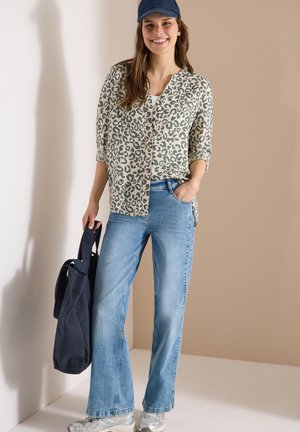 Kobieta uśmiechająca się, ubrana w bluzkę w panterkę, niebieską czapkę, jasnoniebieskie dżinsy, srebrne trampki, trzymająca ciemnoniebieską torbę na ramię, z jedną ręką w kieszeni.