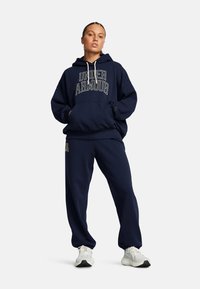 Dunkelblaues Kapuzenoberteil mit dem weißen "Under Armour"-Logo. Verfügt über eine Fronttasche, lockere Passform und passende dunkelblaue Jogginghosen. Weiße Sportschuhe.