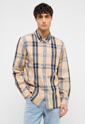 Junger Mann trägt ein beiges, blaues und graues kariertes Hemd mit Kragen und hellblaue Jeans, steht mit einer Hand in der Tasche.