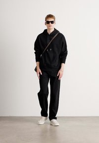 Zwarte hoodie met trekkoorden, bijpassende zwarte broek, witte sneakers en zwarte zonnebril, staand tegen een neutrale achtergrond.