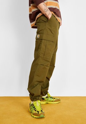 Person iført olivengrønne cargobukser, brun og gul stribet skjorte og neongule og grønne sneakers, stående på gul gulv.