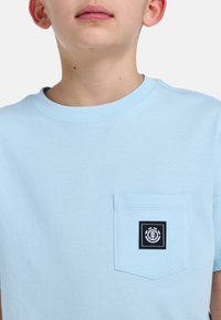 Lysblå bomulds T-shirt med rund halsudskæring, korte ærmer og en venstre brystlomme med et sort emblemmærke.