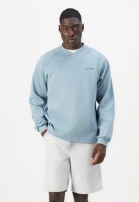 Light blue, long-sleeve sweatshirt with a v-neck design and ribbed cuffs, paired with light gray shorts. Features embroidered logo on chest.