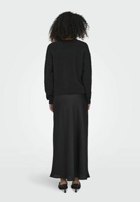 Schwarzer strukturierter Pullover mit überschnittenen Schultern, kombiniert mit einem fließenden schwarzen Maxikleid und abgerundet mit spitzen schwarzen Absätzen.