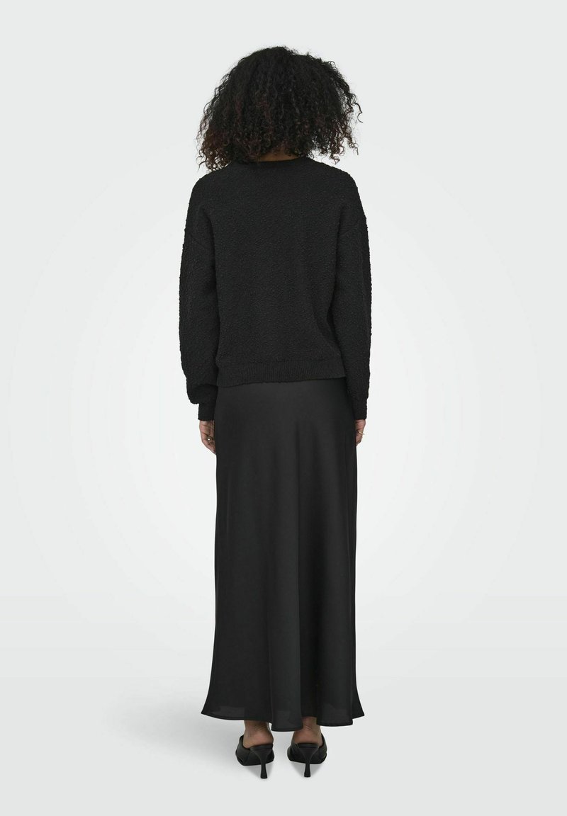 Schwarzer strukturierter Pullover mit überschnittenen Schultern, kombiniert mit einem fließenden schwarzen Maxikleid und abgerundet mit spitzen schwarzen Absätzen.