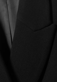 Melnā žakete ar faktūru audumu, ar nošķirtu atloku un vienu krūšu kabatu, izsmalcinātu šuvumu un gludu iekšējo oderi.