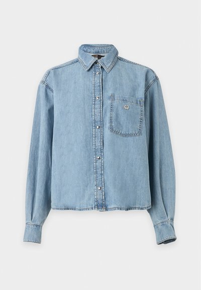 Chemise en denim bleu clair à manches longues, boutonnée, avec un col et une poche poitrine ornée d'un petit détail en forme de cœur.