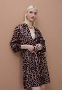 Sukienka w panterkę z miękkiego materiału, o luźnym kroju, zapinana na guziki z przodu, z długimi rękawami i wiązanym w talii paskiem. Złoty naszyjnik i kolczyki.