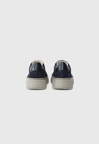 Chaussures en daim marine avec des accents en cuir noir et une semelle en caoutchouc gris épais, présentant un détail tissé autour du bord de la semelle.