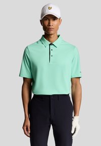 Mintgrön kortärmad polo skjorta med krage och tre knappar, kombinerad med mörka byxor, kompletterad av en vit keps med en gul logotyp.