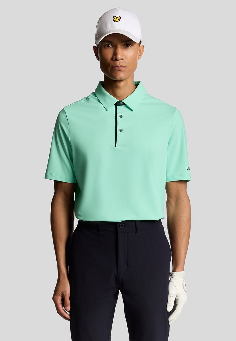 Mintgrön kortärmad polo skjorta med krage och tre knappar, kombinerad med mörka byxor, kompletterad av en vit keps med en gul logotyp.